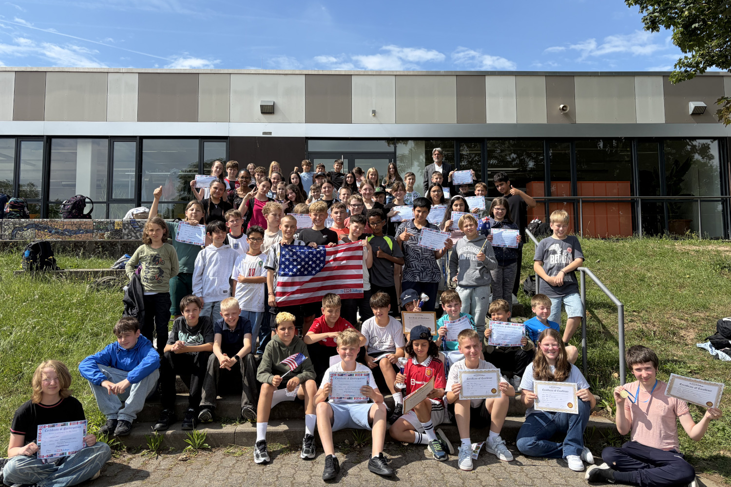 Ein großes Gruppenfoto von etwa 50 Schülerinnen und Schülern der Bertha-von-Suttner-Schule, die stolz auf den Treppenstufen vor dem Schulgebäude posieren. Viele von ihnen halten ihre Urkunden des „The Big Challenge“-Englischwettbewerbs in die Kamera. In der Mitte der Gruppe halten zwei Schüler eine US-amerikanische Flagge hoch, ein weiterer Schüler im Vordergrund hält eine kleine USA-Fahne. Im Hintergrund stehen Lehrkräfte und die moderne Glasfassade des Schulgebäudes ist zu sehen.