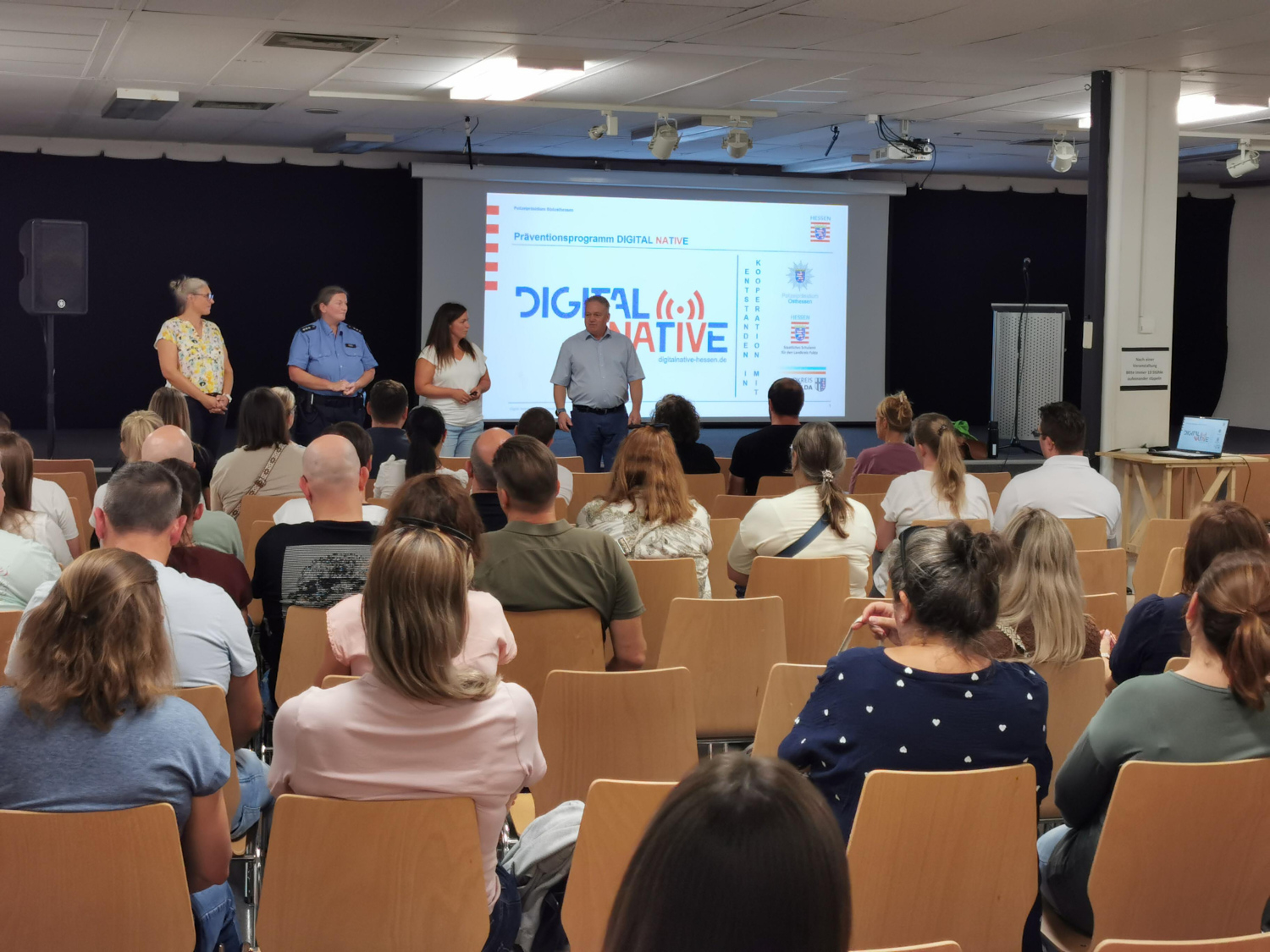 Informationselternabend zum Thema „Digital Natives“ in der Aula der Bertha-von-Suttner-Schule. Vor einer Präsentationsleinwand stehen vier Personen, darunter eine Polizeibeamtin, und sprechen zu den zahlreich anwesenden Eltern und Erziehungsberechtigten.
