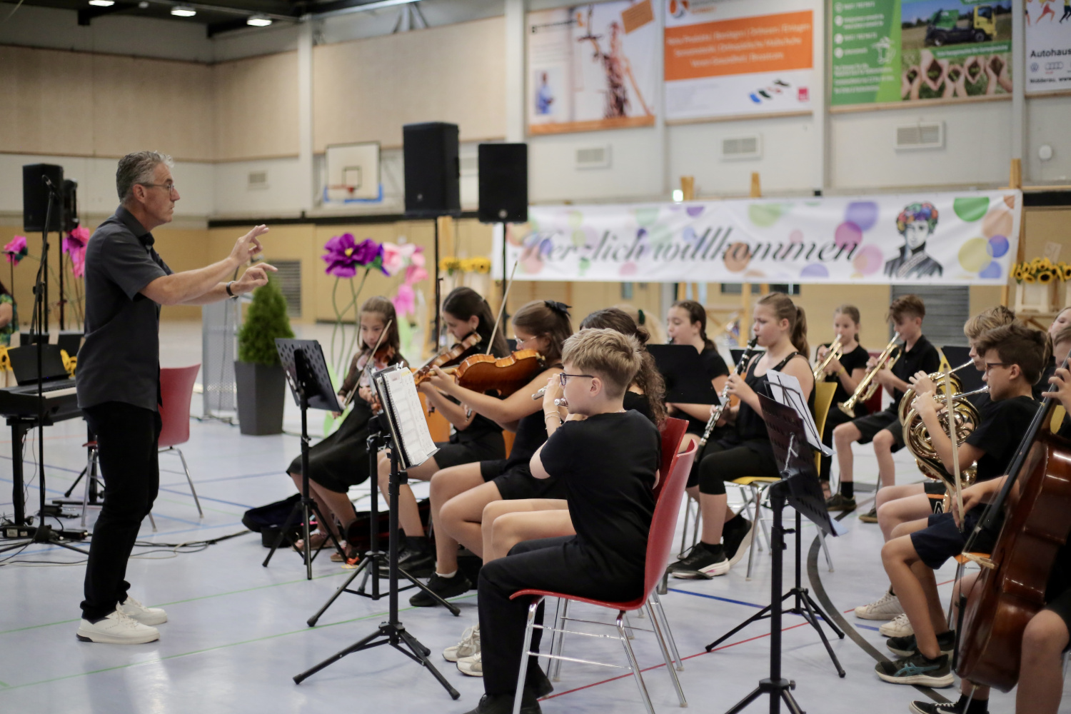 Bei der Einschulungsfeier in der Sporthalle dirigiert ein Musiklehrer die Orchesterklasse. Schülerinnen und Schüler spielen unter anderem Geigen, Klarinetten, Trompeten und Hörner. Im Hintergrund ist ein Willkommensbanner für die neuen Fünftklässlerinnen und Fünftklässler zu sehen.