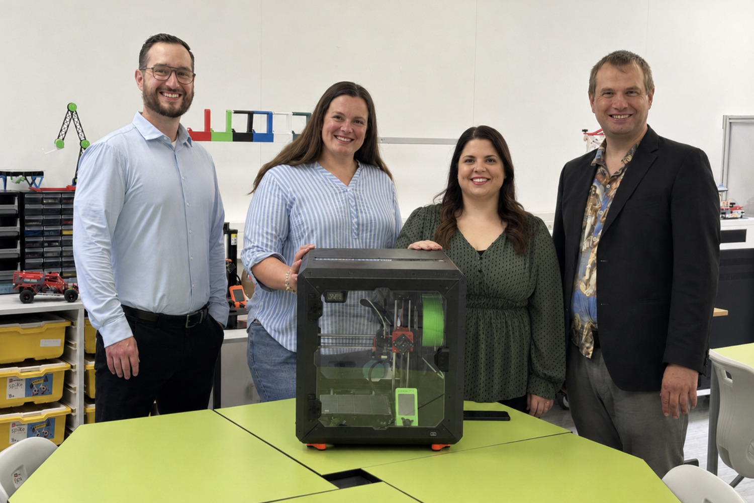 Vier Personen stehen hinter einem neuen 3D-Drucker im MINT-Zentrum der Schule und lächeln in die Kamera. Der Drucker wurde im Rahmen einer Spende an die Schule übergeben und erweitert die technischen Möglichkeiten für den MINT-Unterricht.
