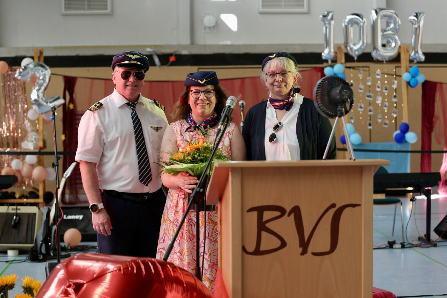 Schulleiter Harald Klose, die ehemalige Schulelternbeiratsvorsitzende Tanja Zelenic und die Stufenleiterin Sybille Michelsen in Piloten- und Flugbegleiter-Uniformen hinter einem Rednerpult mit dem Logo „BVS“ bei der Abschlussfeier der Bertha-von-Suttner-Schule.